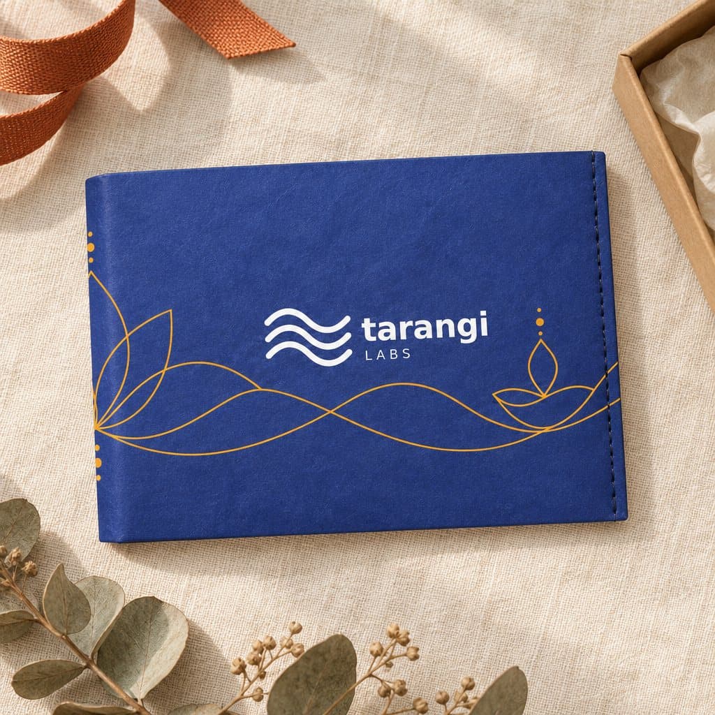 The Tarangi Labs wallet, closed on cream linen beside a ribbon and a gift box: deep indigo, marigold lotus line-work and the logo in white on the front.
